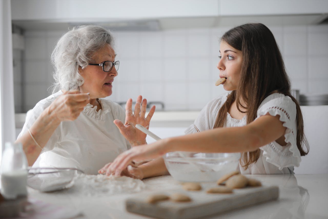 services-03 Calm senior woman and teenage girl in casual clothes looking at each other and talking while eating cookies and cooking pastry in contemporary kitchen at home