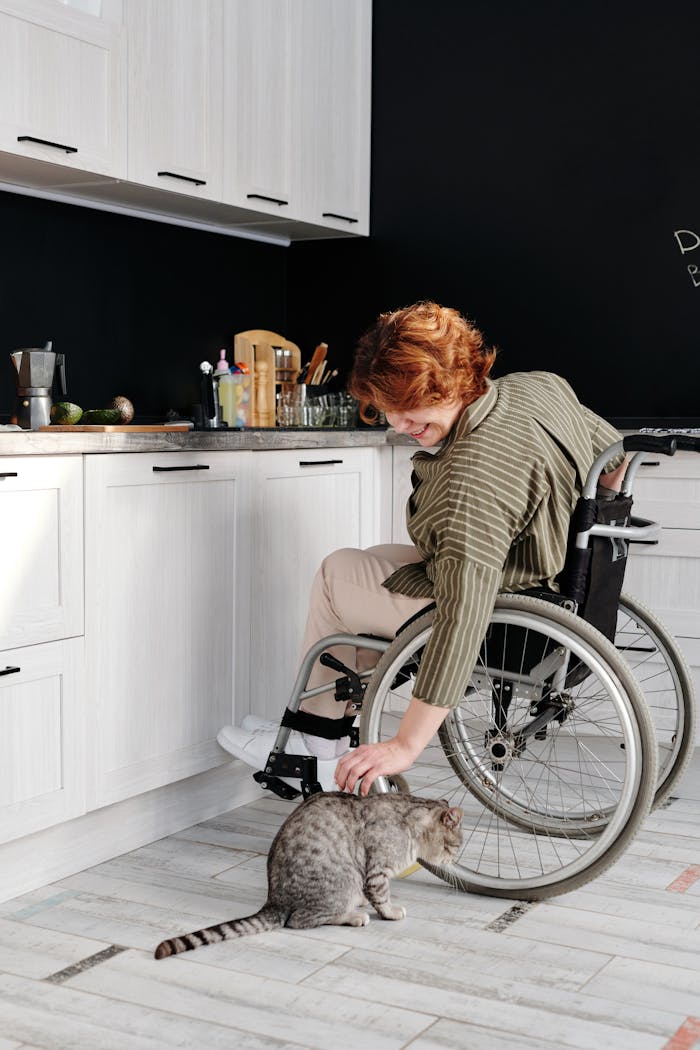 gallery-6 A woman in a wheelchair smiles while petting a tabby cat in a modern kitchen.