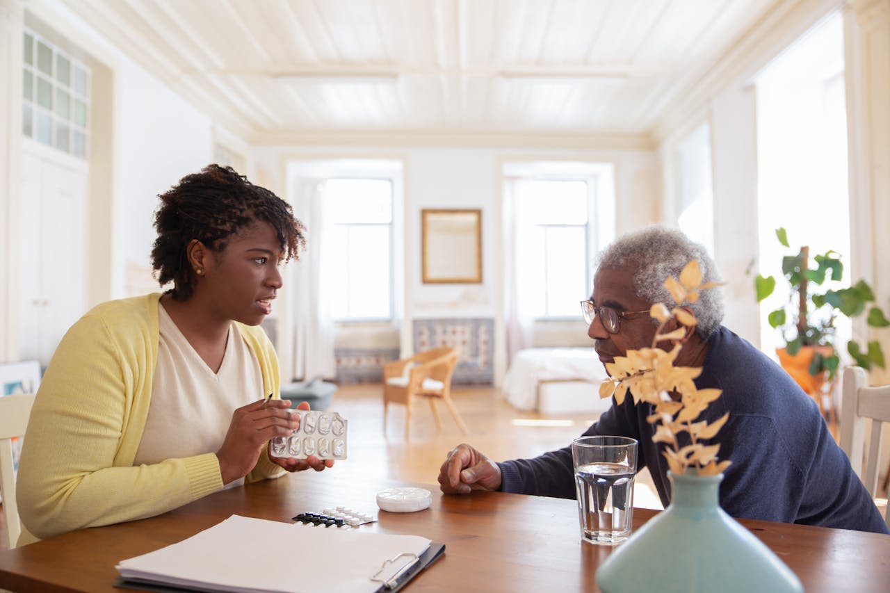 services-02 An elderly man sits with a caregiver discussing medication at a table in a bright room.